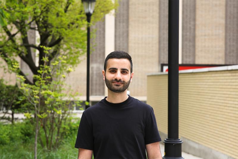 Edmund Shalhoub smiling and standing outdoors in a garden with skyscrapers in the background.
