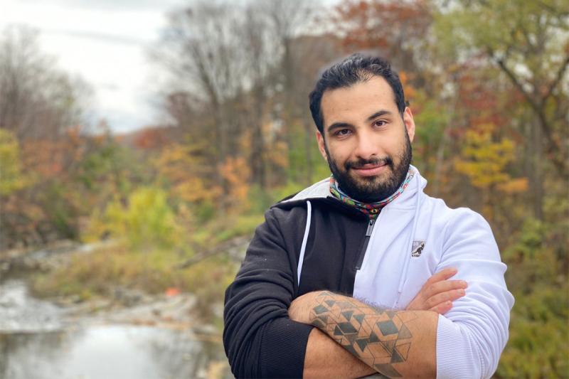Faraz Khoshbakhtian smiles, standing by a river in a wooded area.