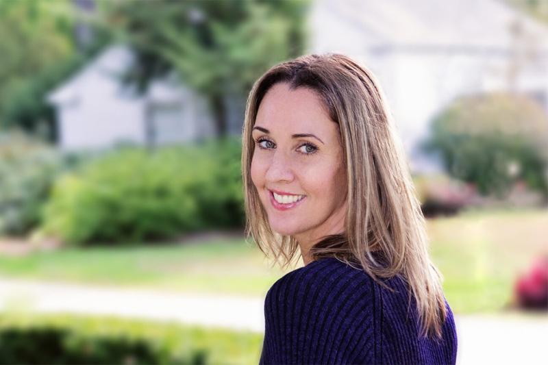 Natalie Jenner looks over her shoulder and smiles. She is standing beside a street in a residential neighbourhood.