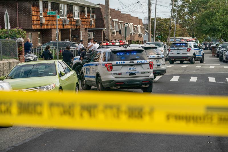 Police tape blocks off a residential street. Behind the line, police gather around their parked cars and talk seriously.