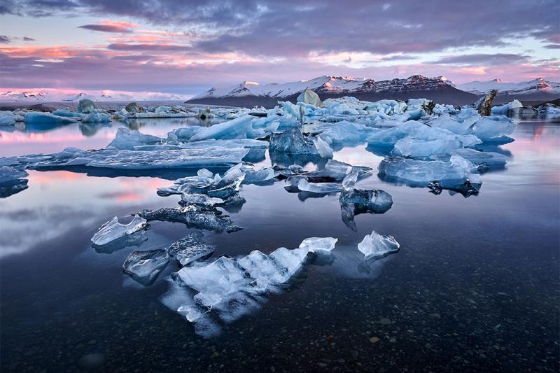 Melting ice floats in a glass-calm Arctic inlet, with snow-covered mountains in the background.
