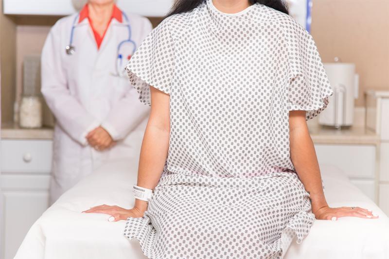 A white woman sits on a hospital bed in a hospital gown, while a figure in a white coat stands behind.
