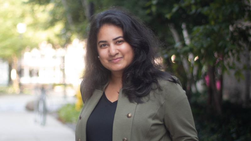 Ishita Aggarwal smiles, standing on a sunny sidewalk outdoors.