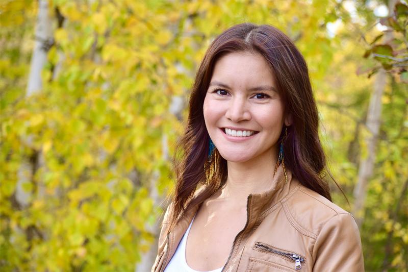 Itoah Scott-Enns smiles, standing outdoors by birch trees.