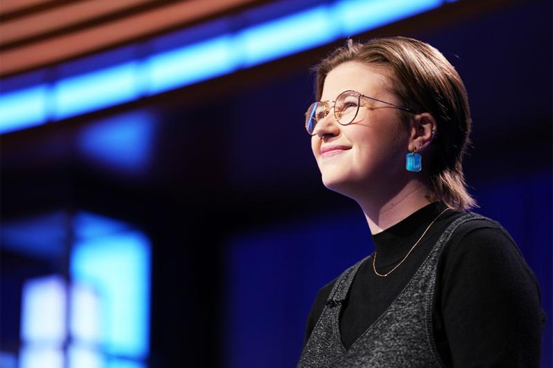 Mattea Roach smiles happily, standing in the lights on the Jeopardy TV set.