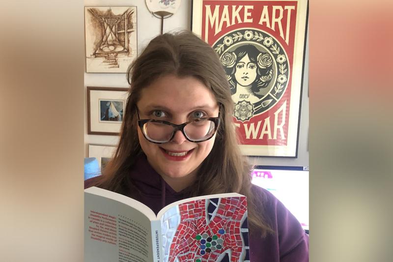Jessica Watkin holds up an open book. Behind her, a poster that says, make art not war.