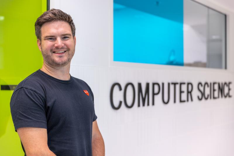 Kent Fenwick smiles for the camera next to a sign that says Computer Science.