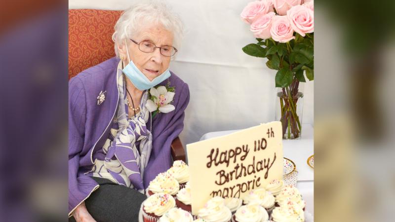 Marjorie Douglas, wearing a flower in her buttonhole, smiles at a tray of cupcakes and a sign saying Happy 110th Birthday.