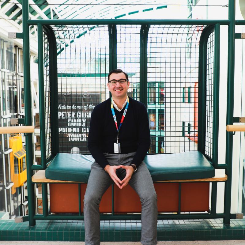 Michael Gritti smiles, as he sits in a balcony alcove with a large atrium behind him.
