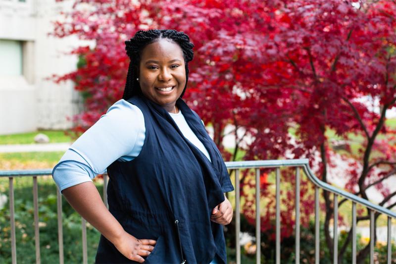 Nicole Mfoafo-M'Carthy smiles as she stands hand on hip in a garden outdoors.
