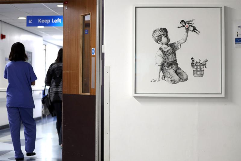 A nurse walks past a hospital artwork showing a small boy playing with 3 superhero toys: batman, spiderman and a nurse.