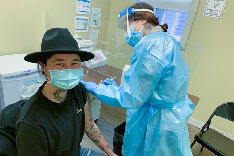 A health-care worker in protective equipment gives a vaccination to a smiling patient in a mask.