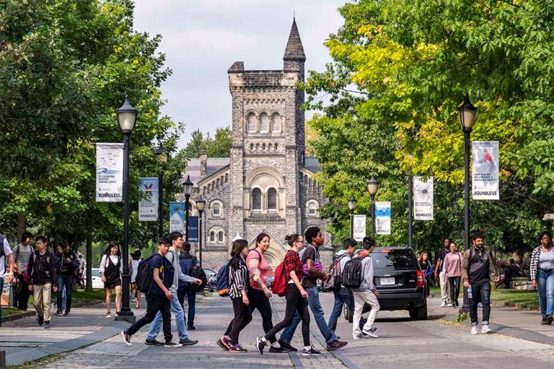 Students wearing backpacks cross the road. University College peeks through trees in the background.