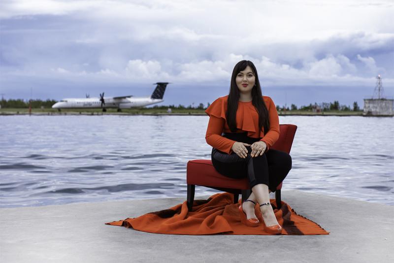Lesley Hampton smiles as she poses in a dramatic ruffled shirt, on a dock across from a lakeside airport.