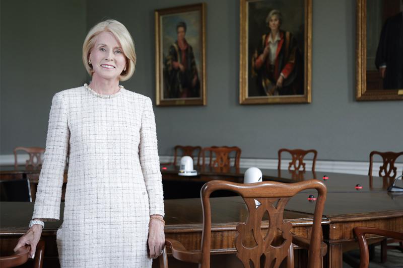 Rose Patten smiles as she stands in front of a large boardroom table, in a room with painted portraits on the wall.