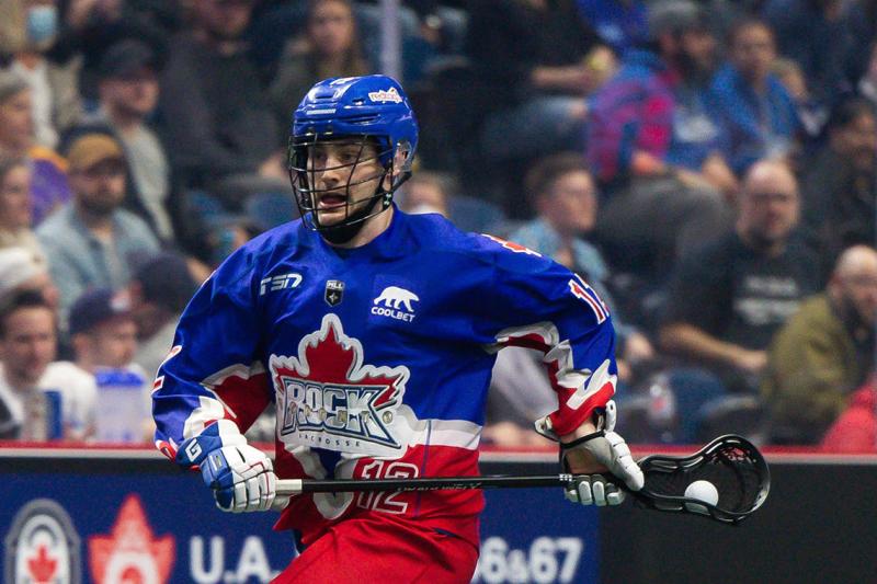 Mitch De Snoo, in the Rock lacrosse team uniform, runs with the ball in his net while a crowd cheers.