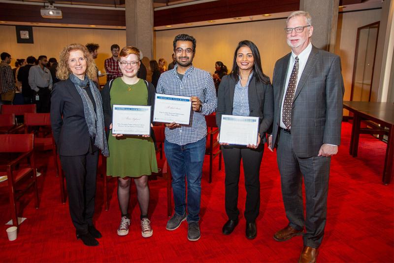 Three students smile and hold up framed awards in a library exhibit room, as a crowd mingles behind them.