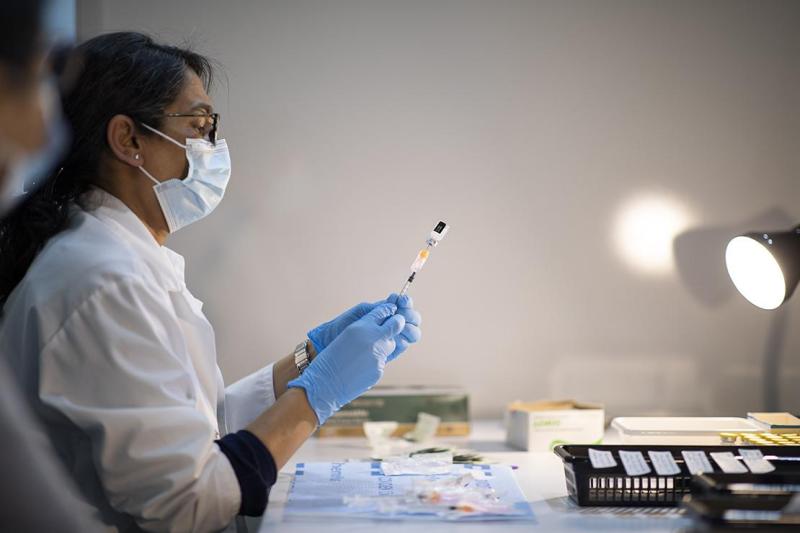 A woman in mask, gown and gloves draws a dose of vaccine into a syringe from a small vial.