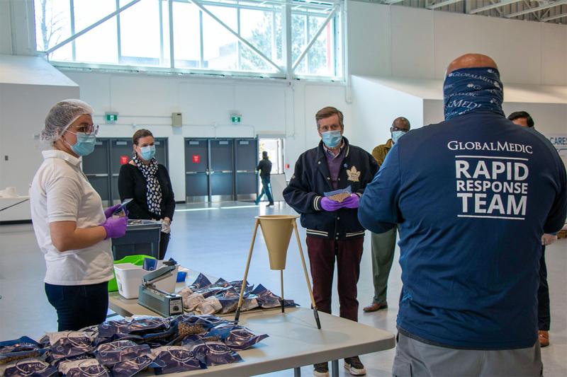 Mayor John Tory, wearing a mask and gloves, listens to a man wearing a hoodie with the words GlobalMedic on it.