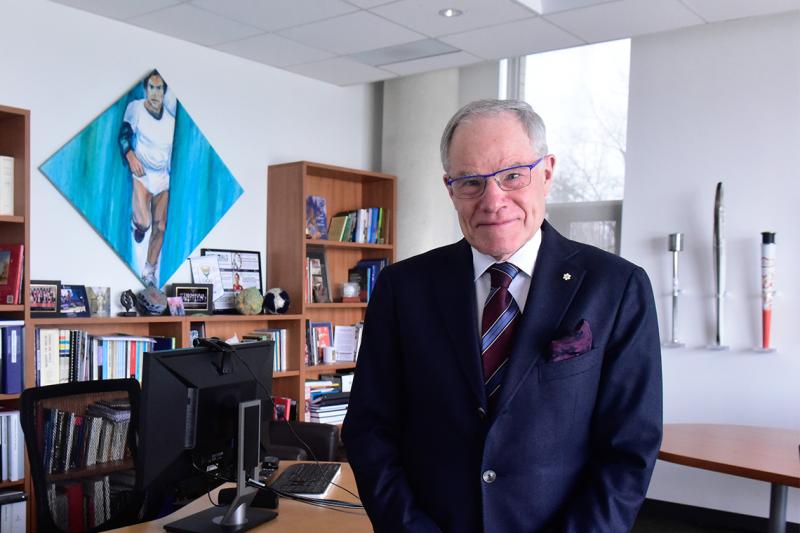 Bruce Kidd smiles as he stands in his office. On the wall are three Olympic torches and a painting of a young Kidd running.