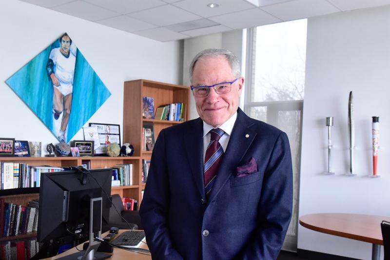 Bruce Kidd smiles as he stands in his office. On the wall are three Olympic torches and a painting of a young Kidd running.