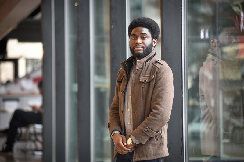 Efosa Obano smiles as he stands outside in front of a glass-walled building.