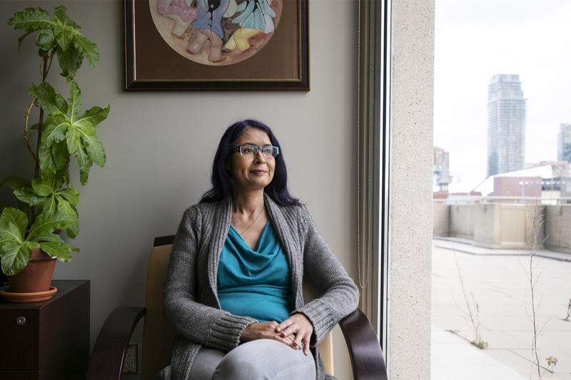 Julie Blair looks thoughtful as she sits by a large window near a painting in an Indigenous style.