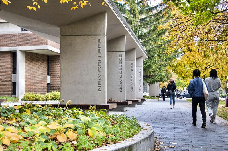 The words New College are engraved in concrete pillars holding up an inviting gateway with benches.