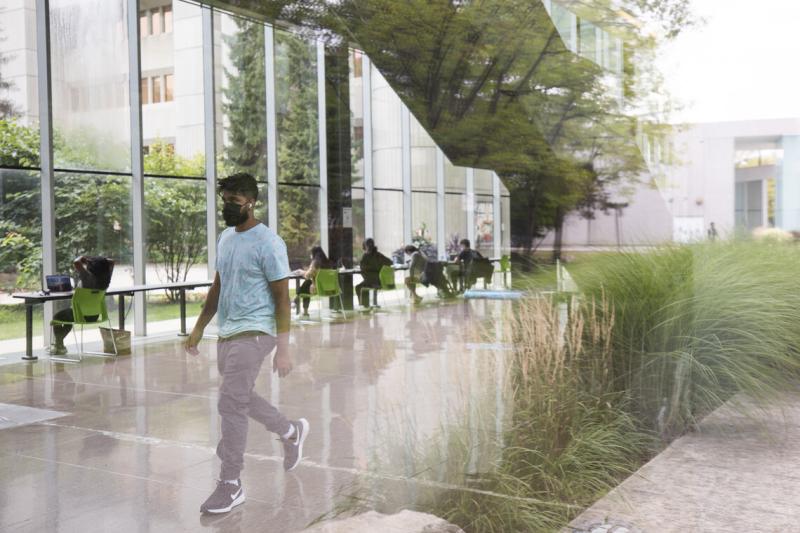 A man wearing a face mask walks past a planter filled with ornamental grasses in a large atrium.