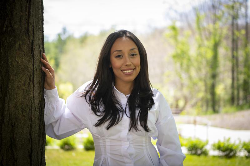 Vanessa Alfaro smiles as she rests her right hand on a tree in a park outside.