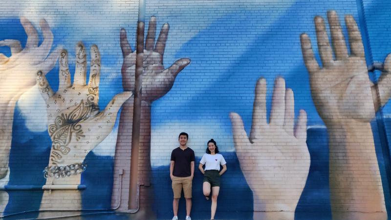 Weiwei Li and Catherine Chan smile as they pose beside a wall mural featuring several giant raised hands.