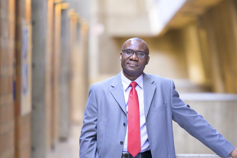 Wisdom Tettey smiles, standing under a skylight in an atrium with one hand on a railing.