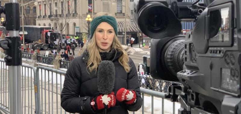Annie Bergeron-Oliver holds a microphone and speaks to a camera in Ottawa, with protesting trucks behind her.