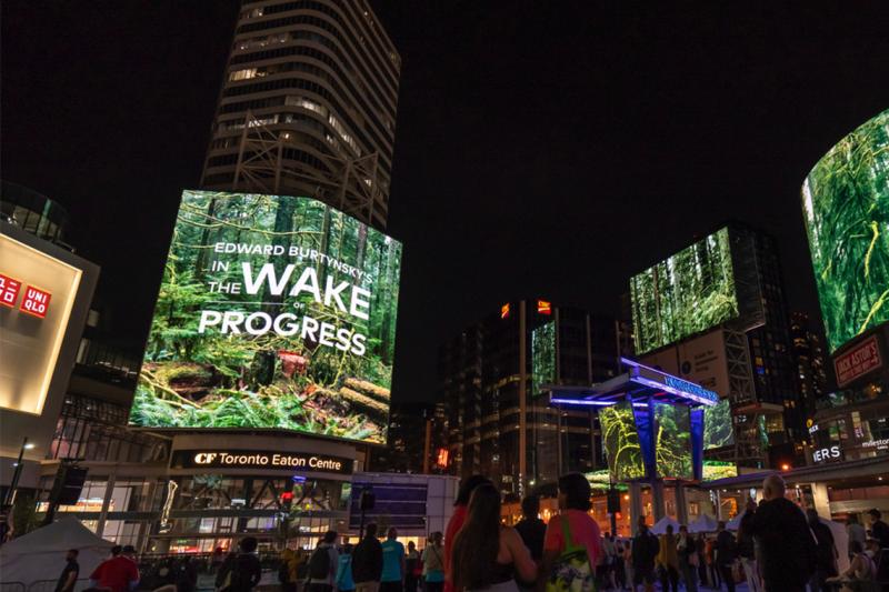 People in a plaza at night look up at a screen. Text reads, Edward Burtynsky's In the Wake of Progress.