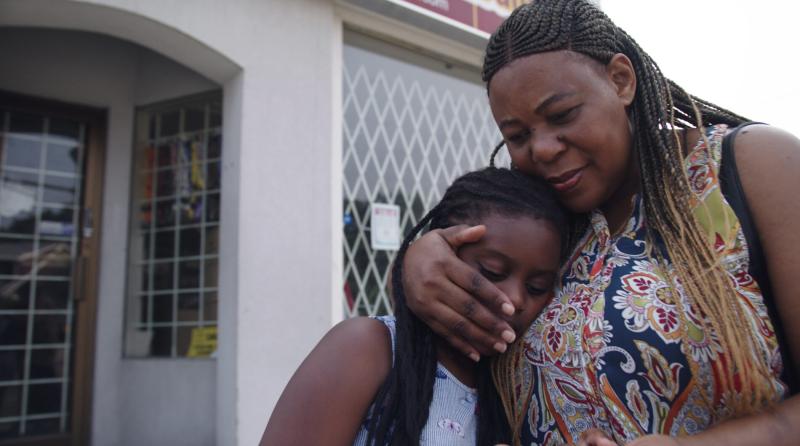 In a still from the film Because She Cares, a woman hugs a little girl who is leaning on her.