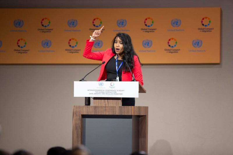 A woman speaking at a podium and gesturing with her right arm.