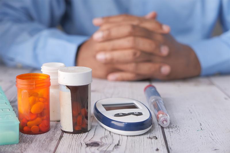 Bottles of pills, a syringe and a monitoring device sit on a table in front of a man's folded hands.