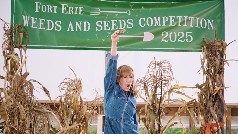 A woman in a denim jumpsuit standing with one arm overhead in front of some dried corn stalks and a sign that reads Fort Erie Weeds and Seeds Competition 2025