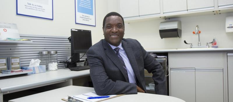 Ian Stewart smiles as he leans one elbow on his desk in a medical office, with a Rx poster on the wall.