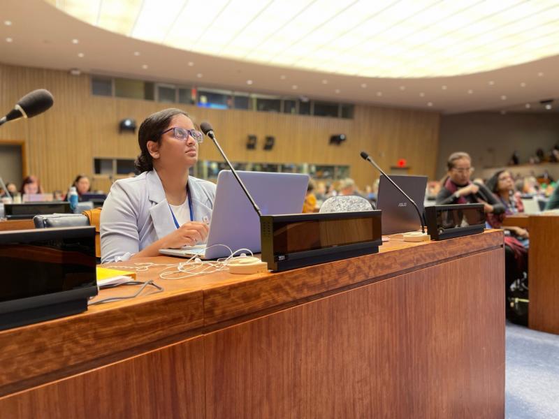 Melanie Ratnam sits at a desk with a microphone in a United Nations assembly room.