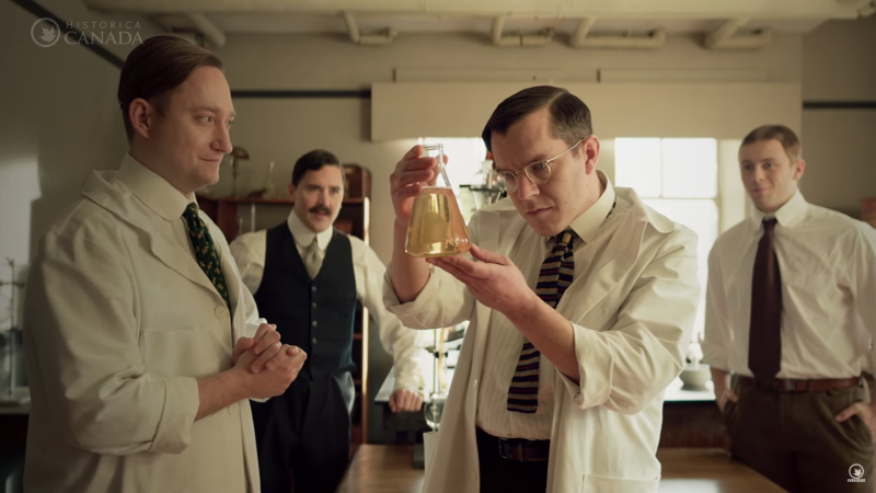 In a screenshot from the Heritage Minute on the discovery of insulin, 4 scientists in 1921 gaze at a beaker of clear liquid.