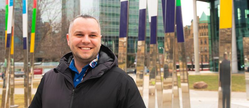 Jaris Swidrovich smiles, standing by an art installation outside the U of T Pharmacy building on College St.