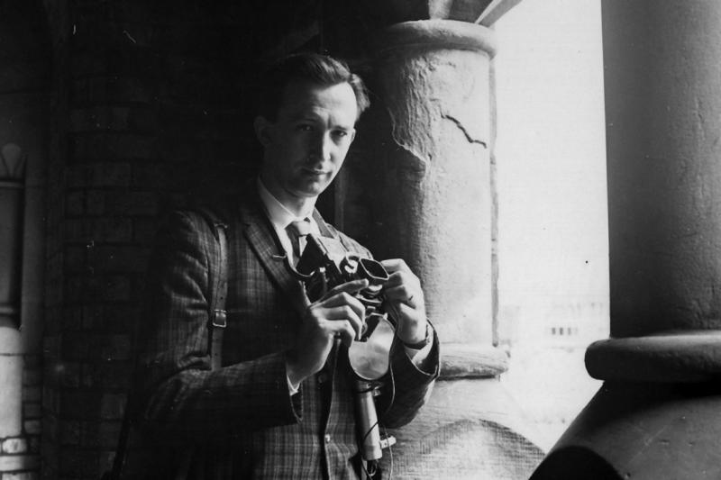 Robert Lansdale stands in shadow on a stone balcony, looking out at the light and holding a camera.