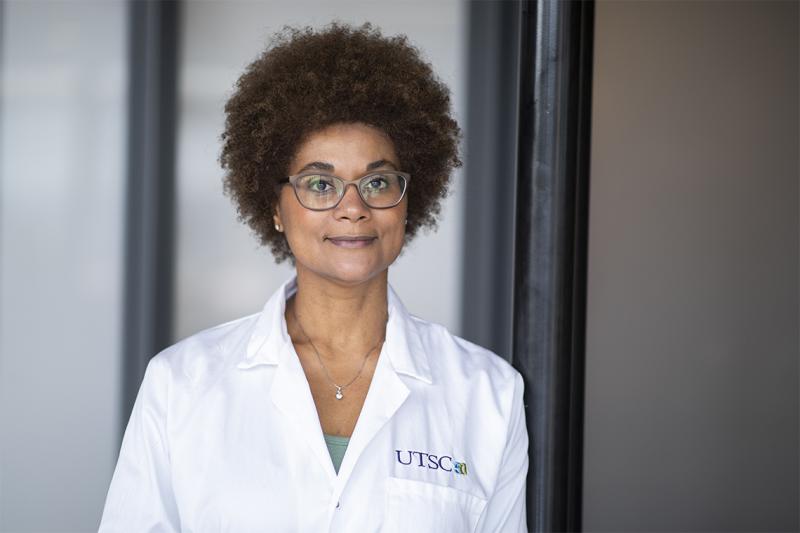 Maydianne Andrare smiles and looks thoughtful, standing in a hallway and wearing a UTSC lab coat.