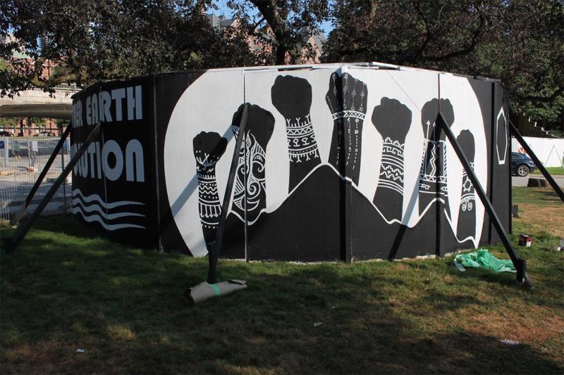 Plywood panels circle a tree. They have been painted with an image of tattooed arms raising their fists in the air.