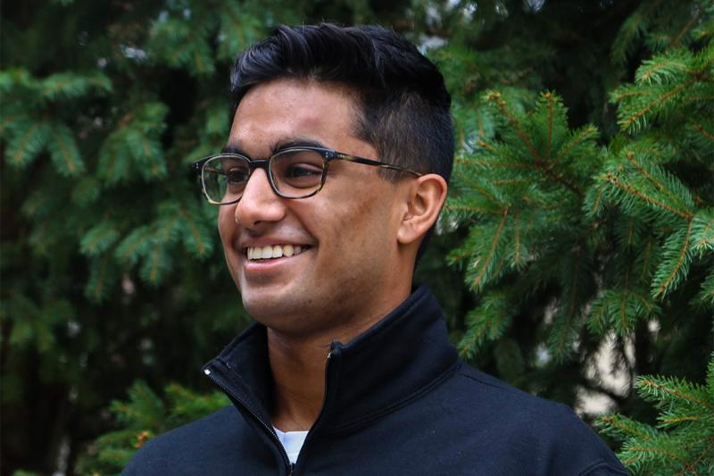 Shrey Jain laughs while standing near fir trees outdoors.