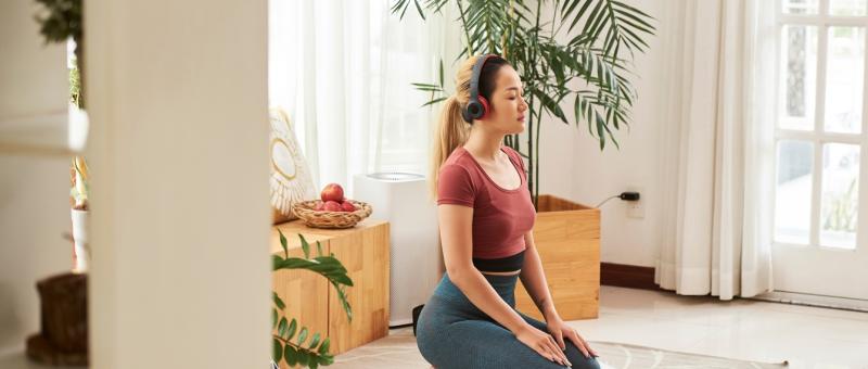 A woman wearing headphones kneels on a yoga mat in a living room.