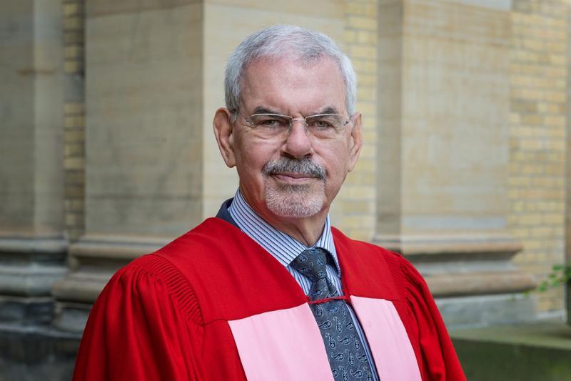 William Troost stands outside Simcoe Hall, wearing academic robes.