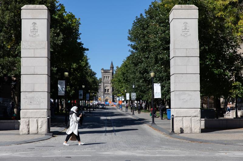 A woman walks past two stone pillars flanking a cobbled street. University College can be seen at the end of the street.