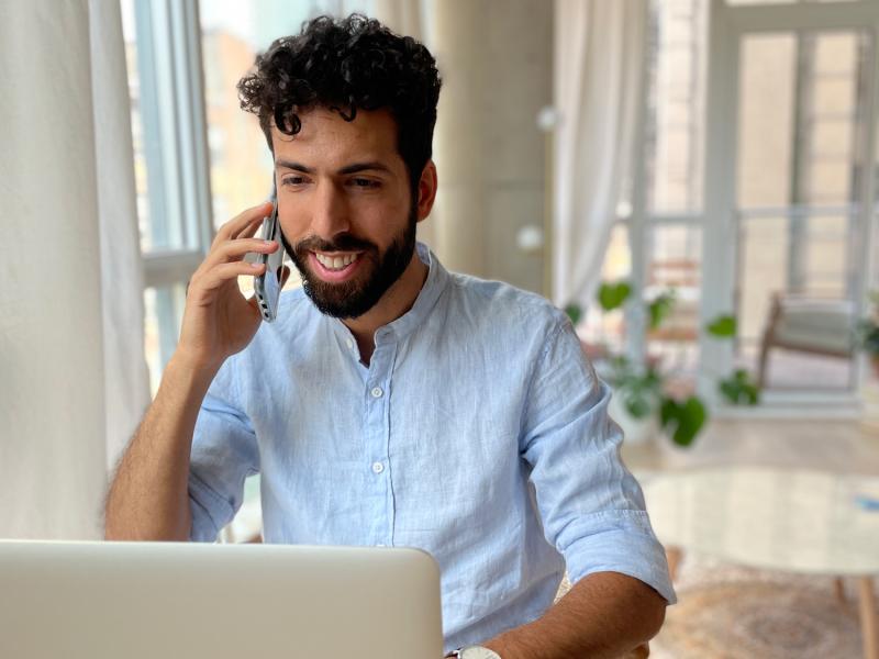 Dr. Latif Murji smiles as he sits at a laptop and talks on a smartphone.